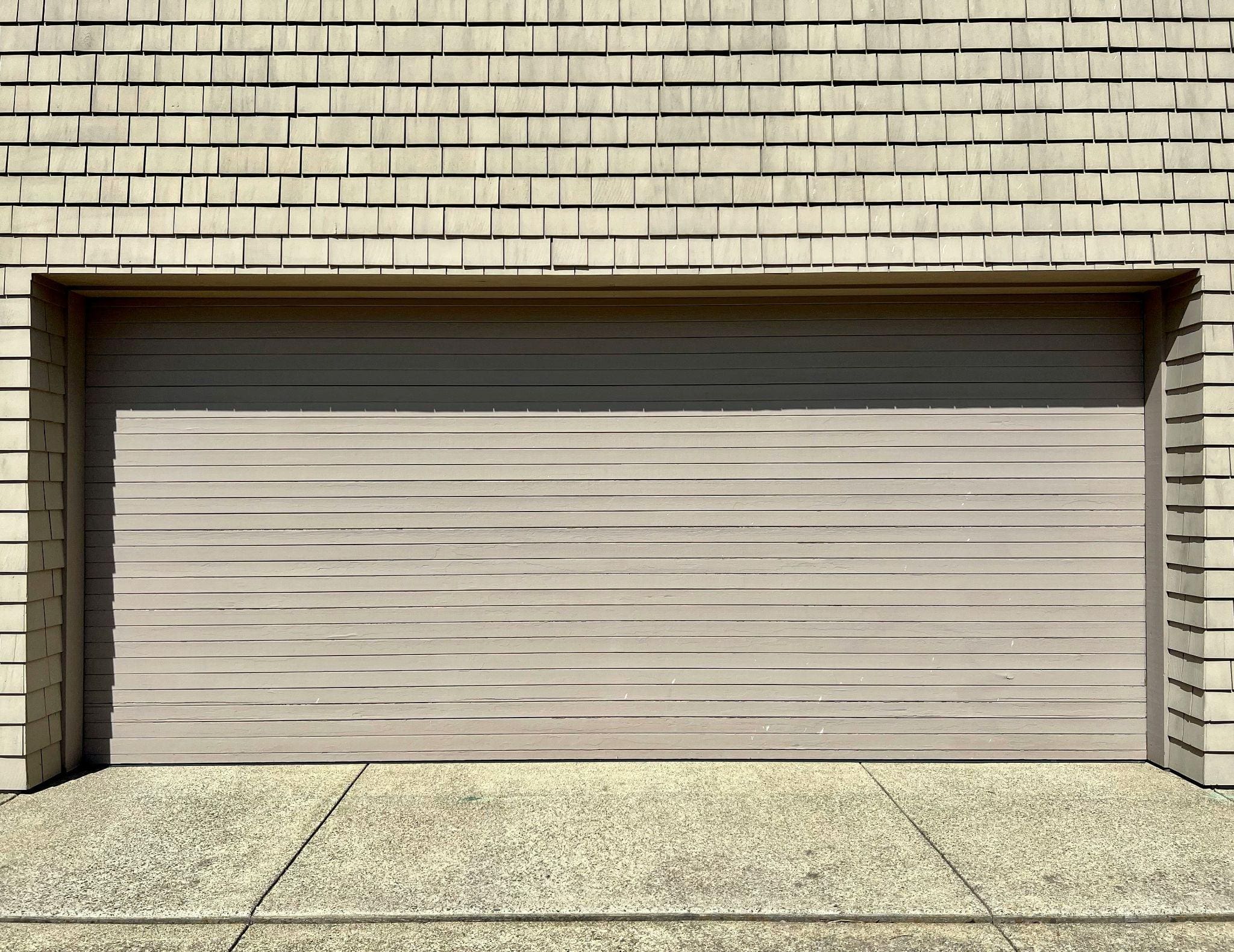 Beige residential garage door with horizontal panels installed beneath light tan exterior shingles and framed by matching siding.